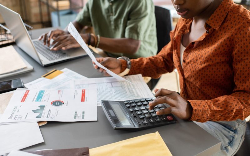 Young African businesswoman or accountant pushing buttons of calculator while working with financial papers against colleague